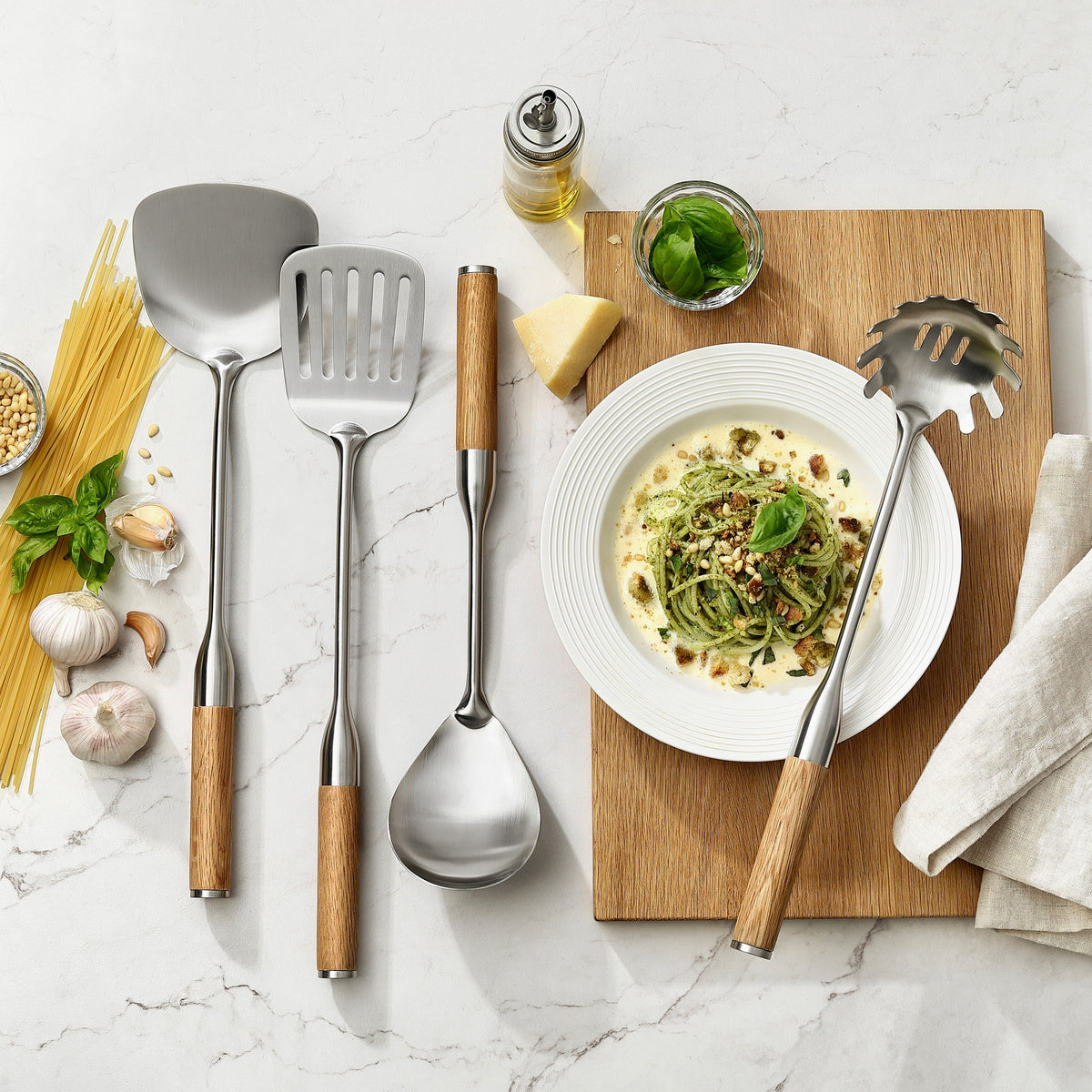 Pfannenwender mit und ohne Schlitze, Pasta- und Servierlöffel mit Griffen aus Eiche neben einem Teller Pasta mit Pesto