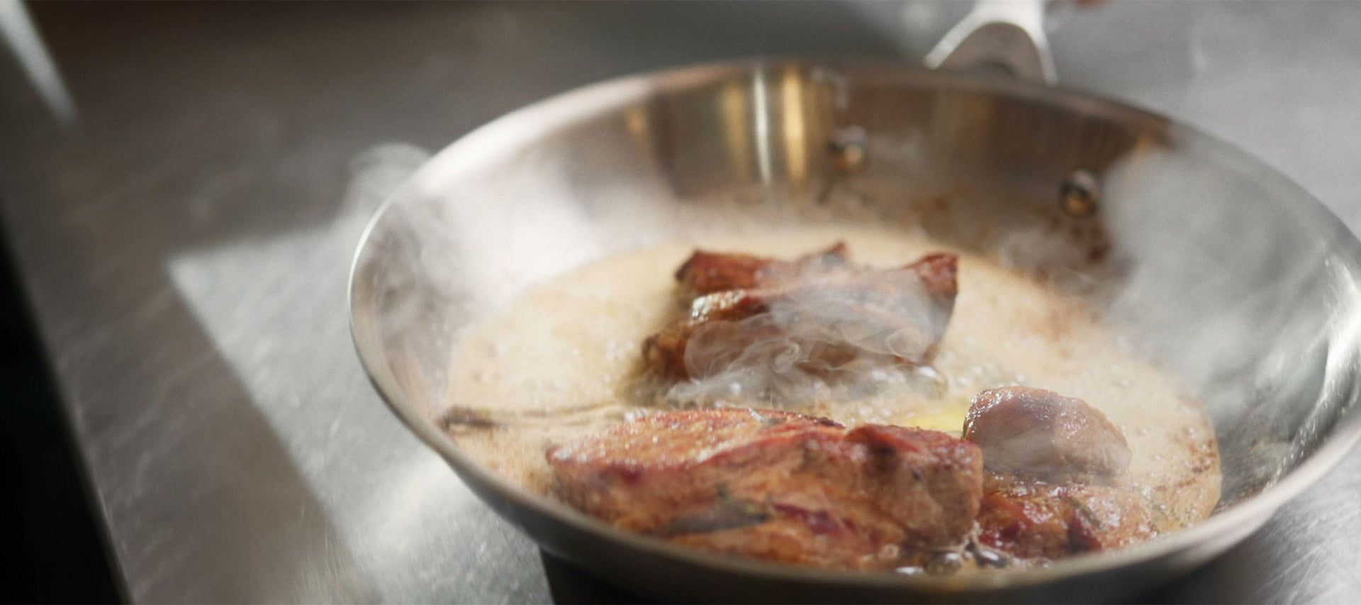Eine Bratpfanne aus Edelstahl, in der Fleisch in schäumender Butter brät, während Dampf aufsteigt