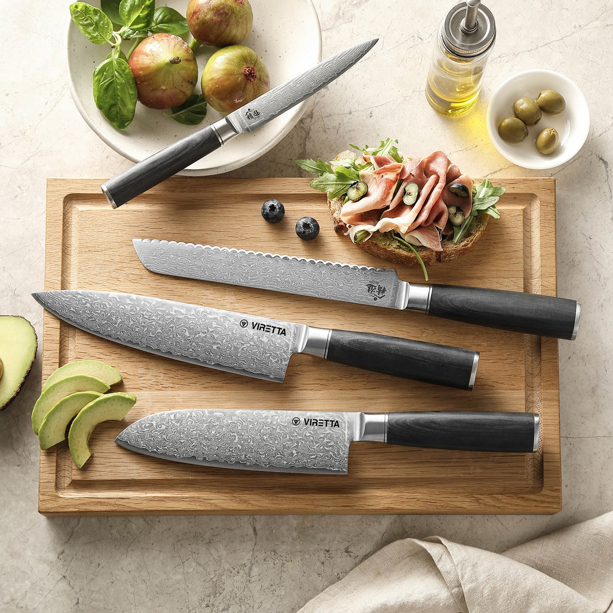 Shōjin Suna Brotmesser, Kochmesser, Santokumesser und Universalmesser auf einem Brett aus Eiche mit Avocado und Oliven