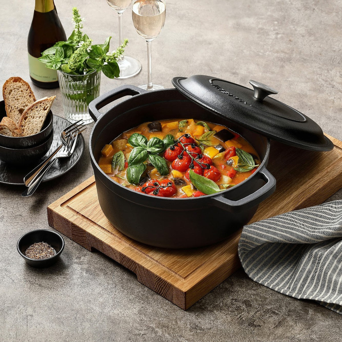 Schieferschwarzer Gusseisentopf mit Ratatouille und schräg aufgelegtem Deckel, auf einem Schneidebrett