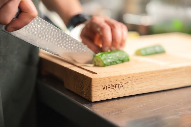 Ein Messer mit gehämmerter Klinge schneidet Gurke auf einem Schneidebrett aus Eiche. Das Brett trägt das Viretta-Logo
