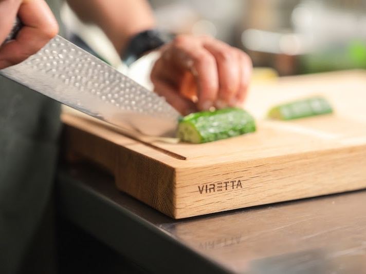 Ein Messer mit gehämmerter Klinge schneidet Gurke auf einem Schneidebrett aus Eiche. Das Brett trägt das Viretta-Logo