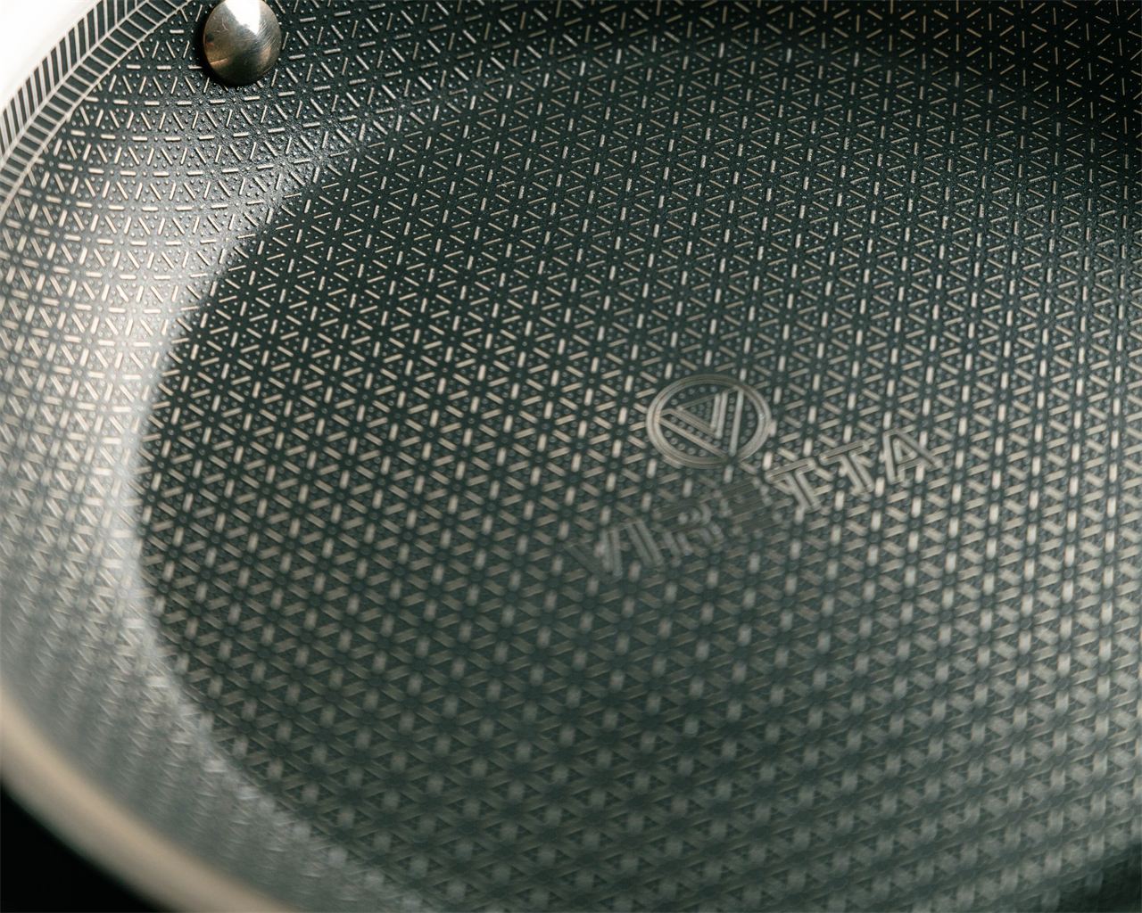 Die Bratfläche einer Hybrid-Pfanne ganz nah: ein Muster aus Stahl über der dunklen Beschichtung, mit dem Viretta-Logo in der Mitte