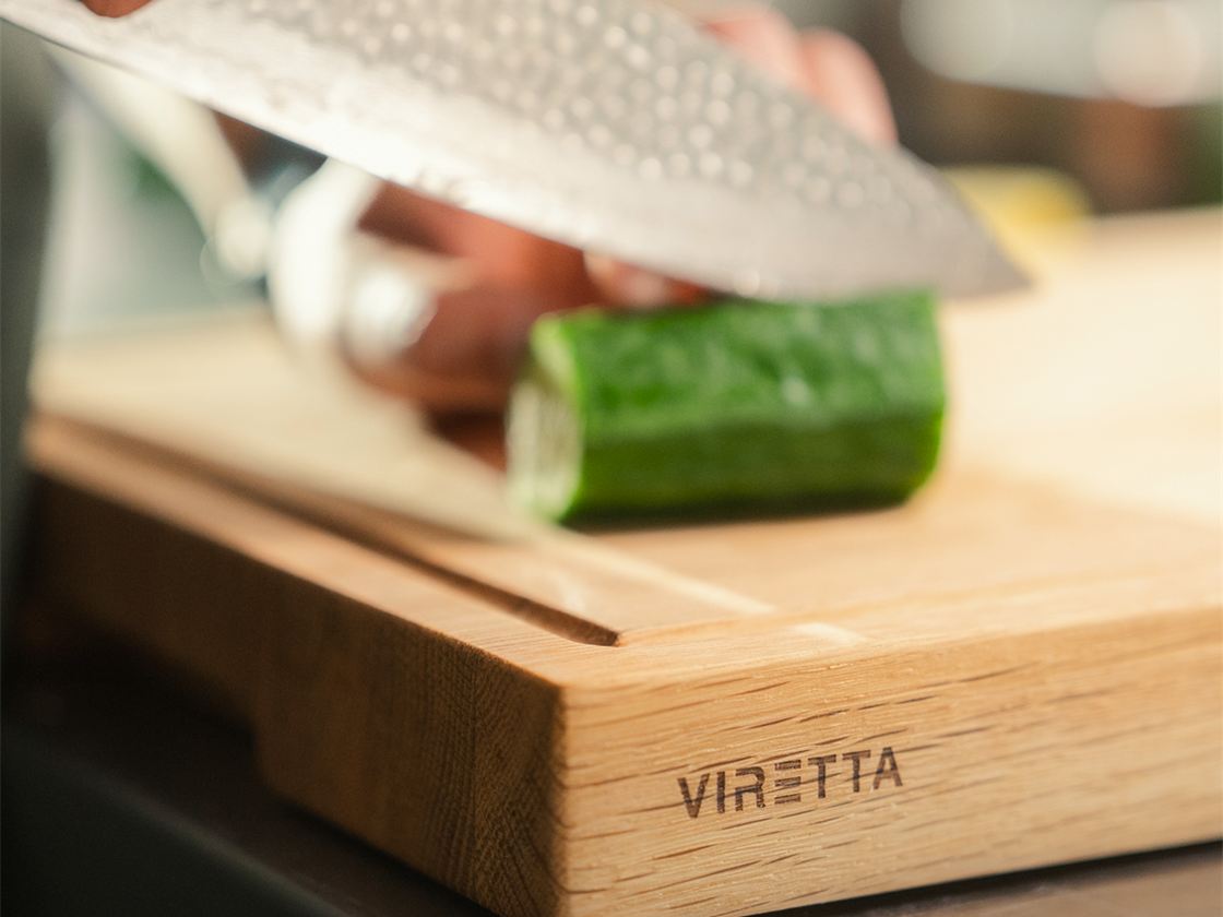Ein Messer schneidet Gurke auf einem Holzbrett. Das Brett trägt das Viretta-Logo
